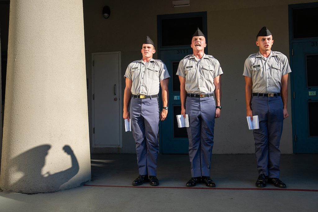 Three uniformed personnel stand in formation outdoors, one with an open mouth, holding papers, under bright sunlight casting a shadow.