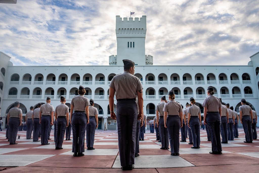 Uniformed personnel stand at attention in formation on a checkered courtyard before a white building with a castle-like tower.