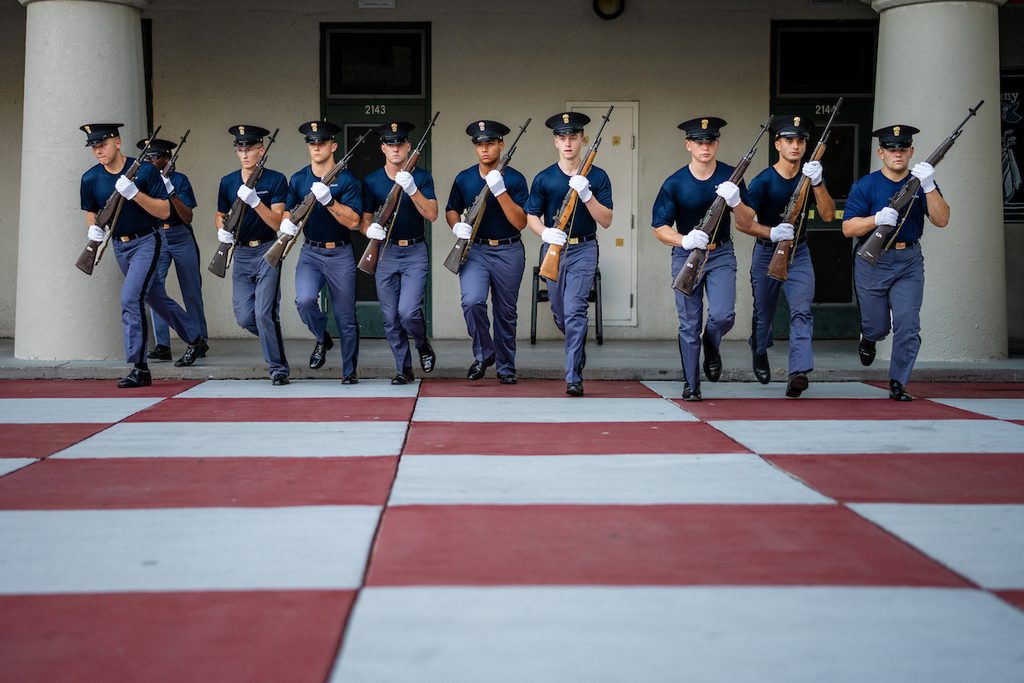 A line of uniformed personnel marches in formation across a red and white checkered floor, carrying rifles and wearing white gloves.