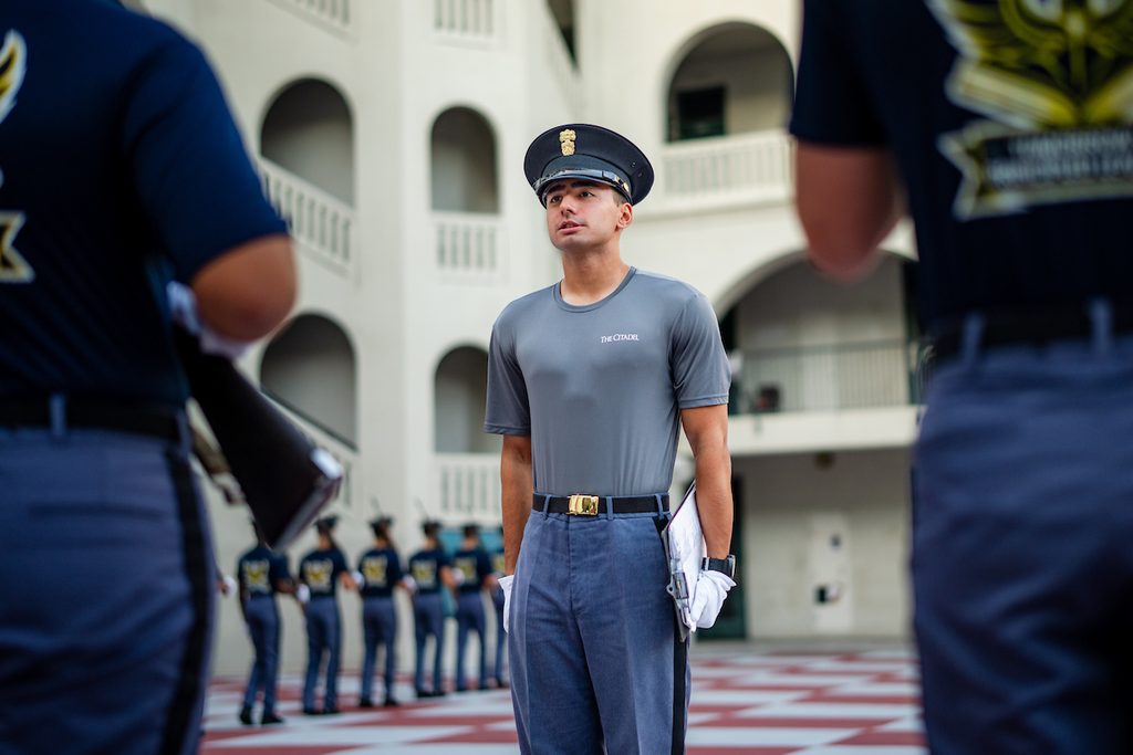 A uniformed individual stands at attention outdoors, holding papers, with others in similar attire visible in the background.