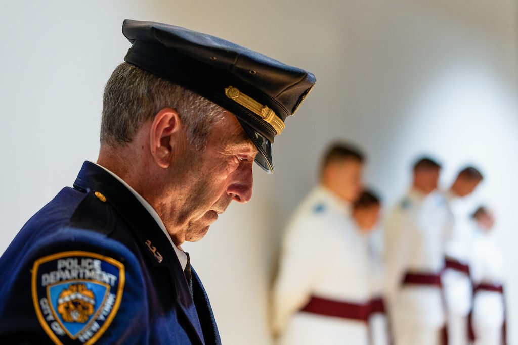 A police officer in uniform stands solemnly with his head bowed, while other uniformed personnel are visible in the blurred background.