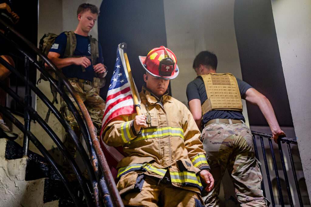 A firefighter in full gear holds an American flag while ascending a staircase, accompanied by others in tactical and military-style attire.