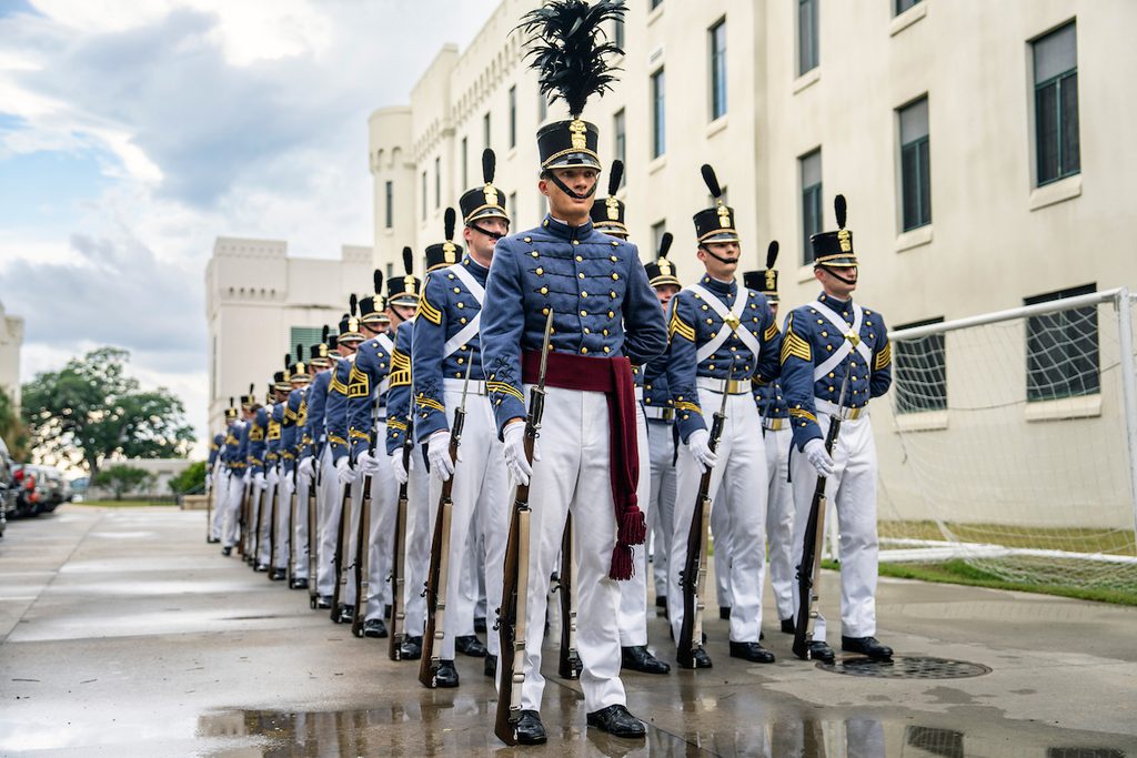 A line of uniformed personnel in formal military attire stand at attention outdoors, holding rifles, with a large building and a soccer goal in the background.