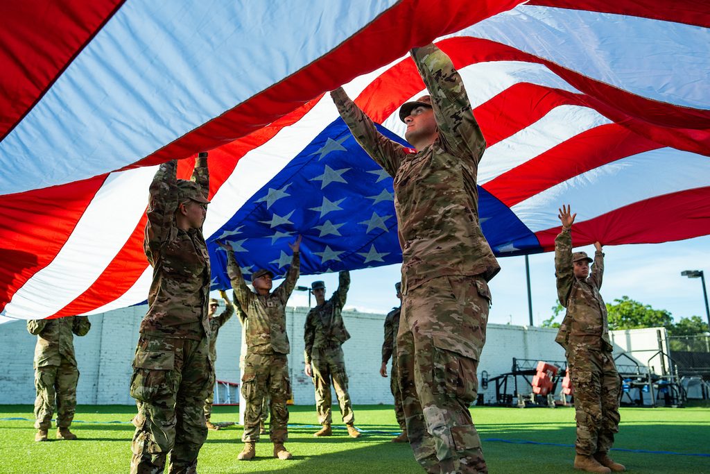 Soldiers in camouflage uniforms hold up a large American flag on a green field under a clear sky.