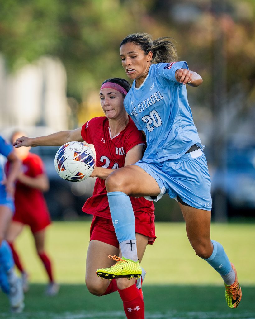 Two soccer players compete for the ball during a match, one in a red uniform and the other in a light blue uniform.