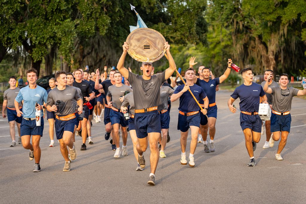 A group of individuals run down a tree-lined road, with one person prominently carrying a large shield above their head.