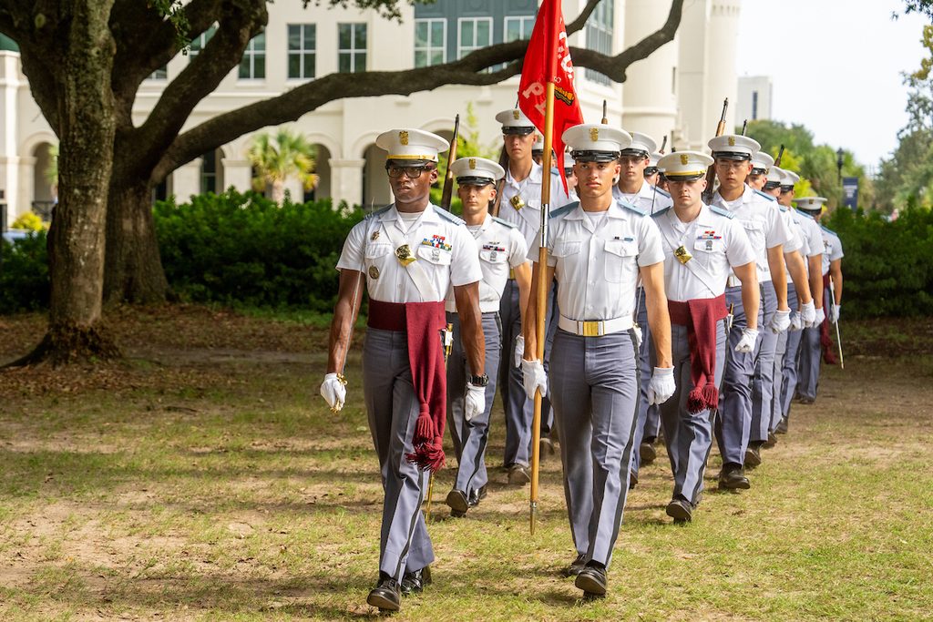 A uniformed military unit marches in formation outdoors, carrying a flag and wearing ceremonial attire.