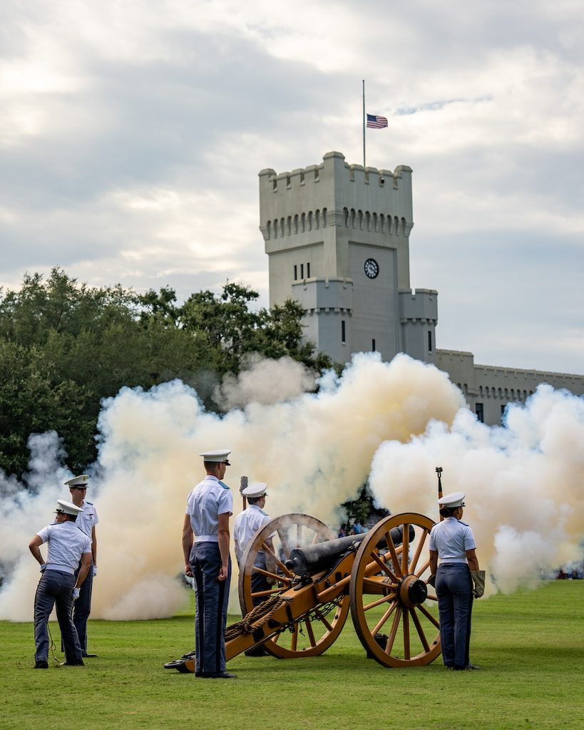 Uniformed personnel stand around a cannon firing, with thick smoke billowing, in front of a castle-like building flying an American flag.