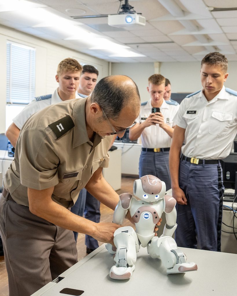 A uniformed individual adjusts a small humanoid robot on a table while several others observe in a classroom setting.