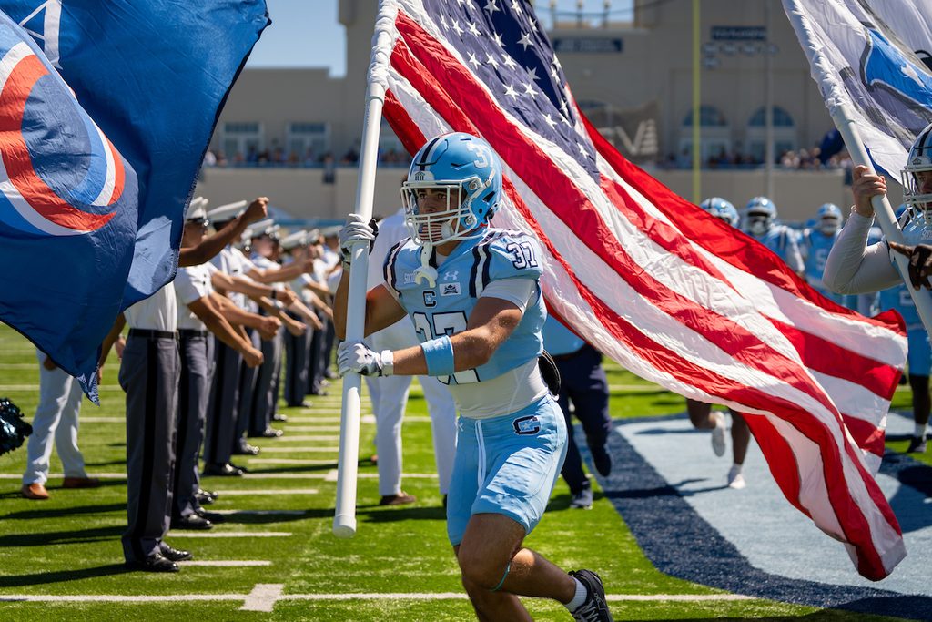 An athlete in a light blue uniform carries a large American flag during a football game entrance.