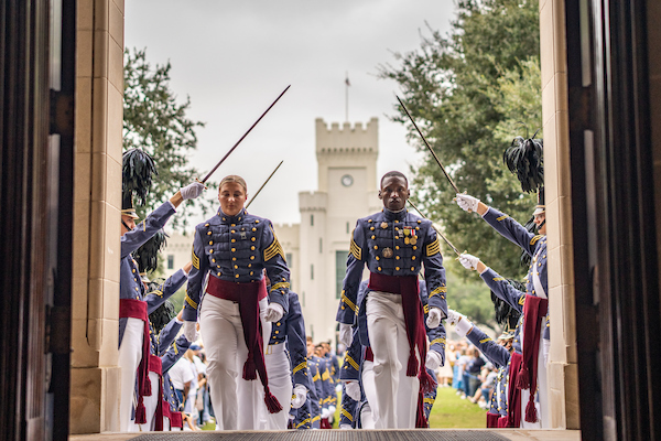 Uniformed cadets in ceremonial dress march in formation through an archway, carrying rifles, with a historic tower visible in the background.