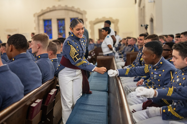 A cadet in uniform smiles as she extends her hand to shake with another cadet seated in a church pew.