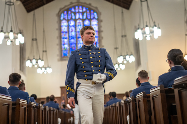 A cadet in a formal military uniform walks down the aisle of a church, holding a white object, with other uniformed individuals seated in wooden pews.