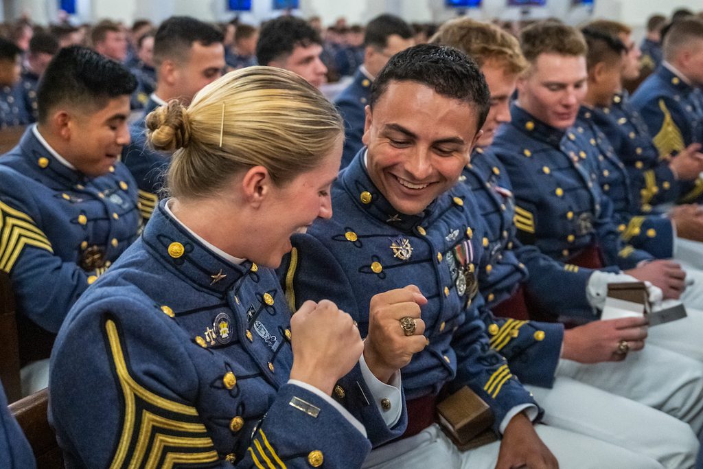 Uniformed personnel in blue dress attire sit in rows, smiling and laughing together during an event.