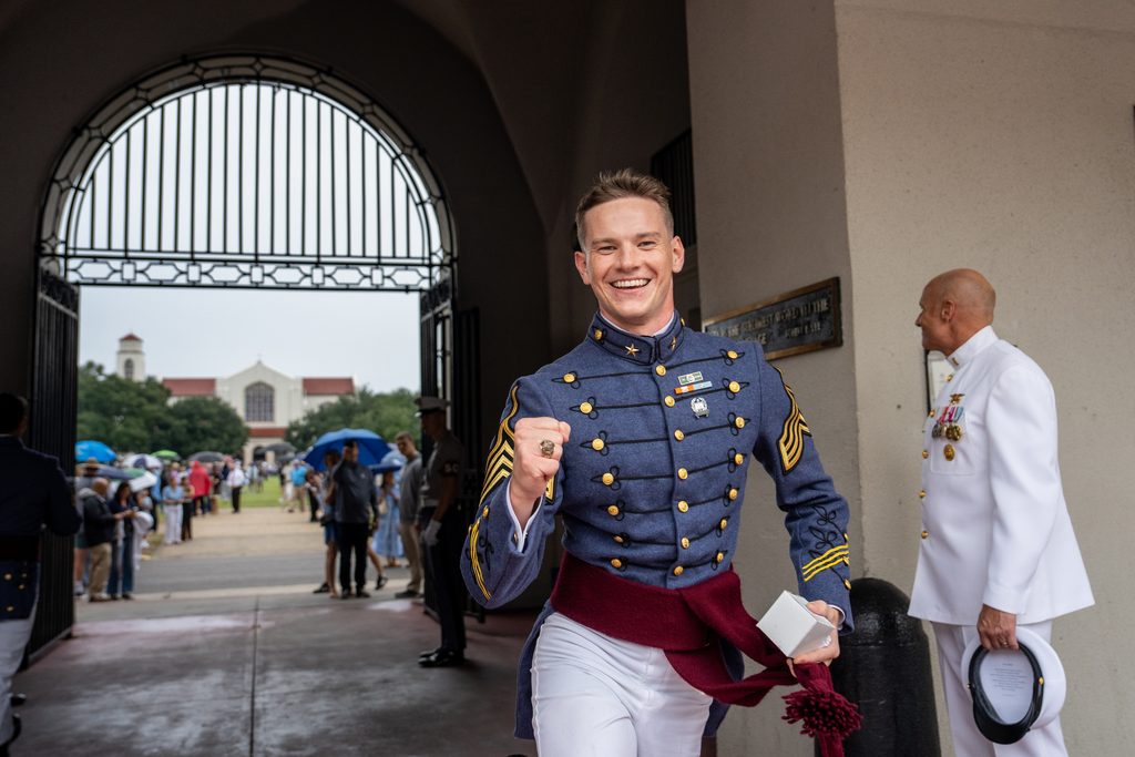 A cadet in a formal military uniform smiles broadly while holding a ring and a box, standing under an arched entrance with others in the background.