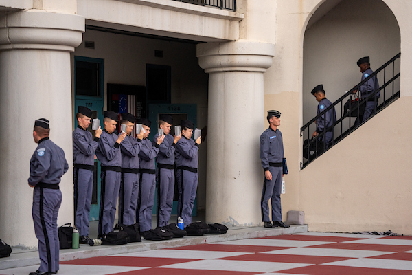 Uniformed personnel stand in formation outside a building, some holding tablets, while others descend stairs.