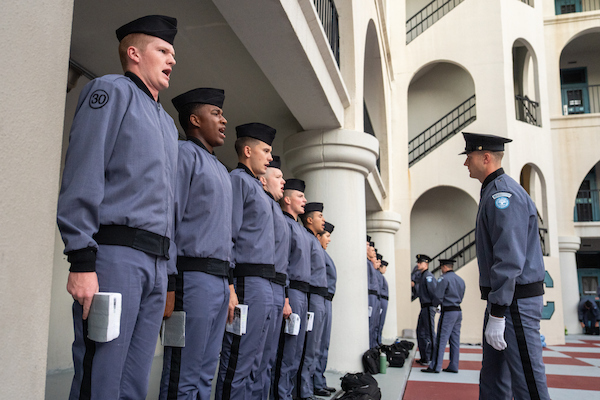 Uniformed personnel stand in formation outdoors, singing or reciting under the supervision of an officer.