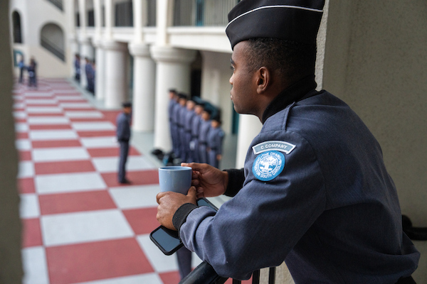A uniformed personnel member from C Company, South Carolina, stands on a balcony holding a mug and a phone, observing a line of similarly dressed individuals in a courtyard with a red-and-white checkered floor.
