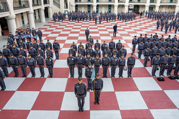A large group of uniformed personnel stands in precise formation on a red and white checkered courtyard, with a few individuals positioned at the front.