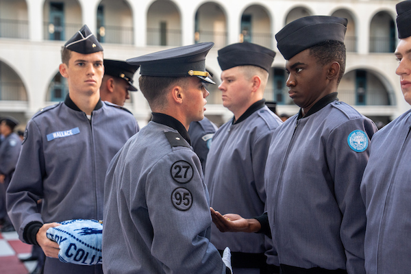 Uniformed personnel in gray jackets and caps stand in formation, with one individual holding a blue cloth item.