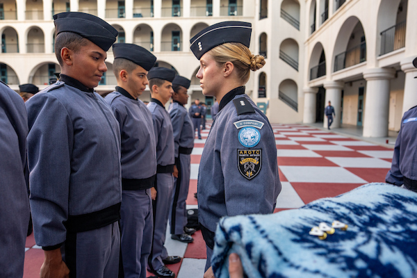 A line of uniformed personnel stands at attention in a courtyard with arched buildings, while one individual in the foreground holds a patterned blanket.