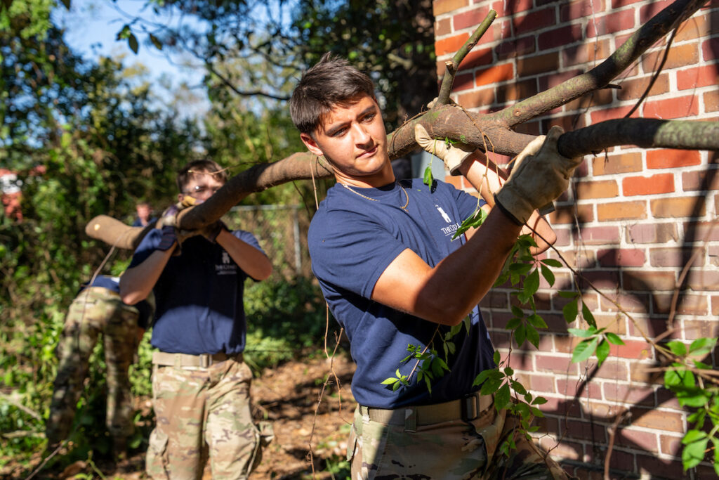 Two individuals carry a large branch outdoors, with one in the foreground and another slightly behind, near a brick wall.