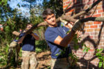 Two individuals carry a large branch outdoors, with one in the foreground and another slightly behind, near a brick wall.