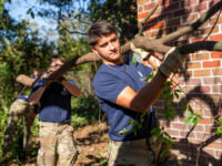 Two individuals carry a large branch outdoors, with one in the foreground and another slightly behind, near a brick wall.