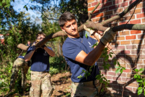 Two individuals carry a large branch outdoors, with one in the foreground and another slightly behind, near a brick wall.