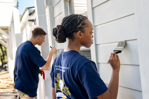 Two individuals paint the siding of a house while wearing matching blue shirts.