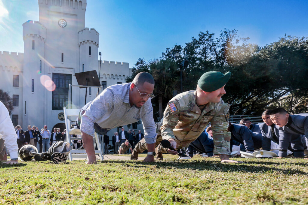 A man in a button-down shirt and a soldier in uniform perform push-ups on a lawn in front of a castle-like building while a group of people watch.