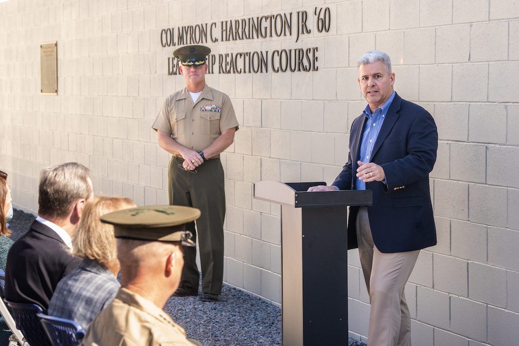 A person in civilian attire speaks at a podium while a military officer stands at attention, with a plaque honoring Col. Myron C. Harrington Jr. '60 visible on a brick wall behind them.