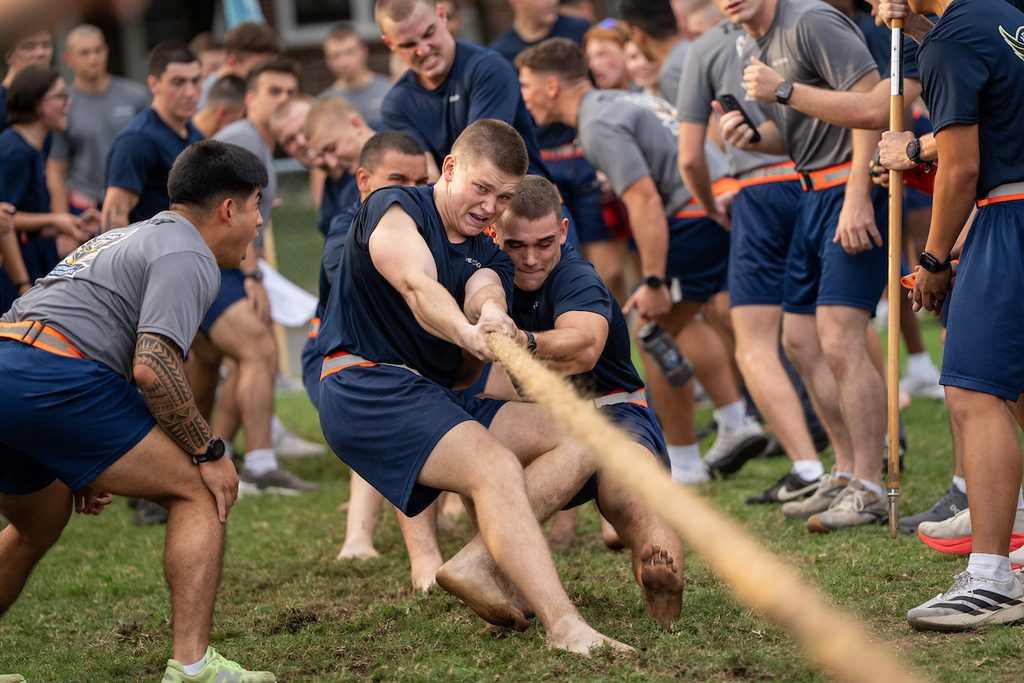 A group of individuals in athletic attire are intensely engaged in a tug-of-war match on a grassy field, with teammates pulling together and spectators watching closely.